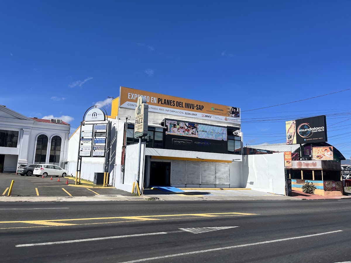 Fachada real del local: edificio blanco de dos plantas con rótulo naranja Experto en planes del INVU SAP
